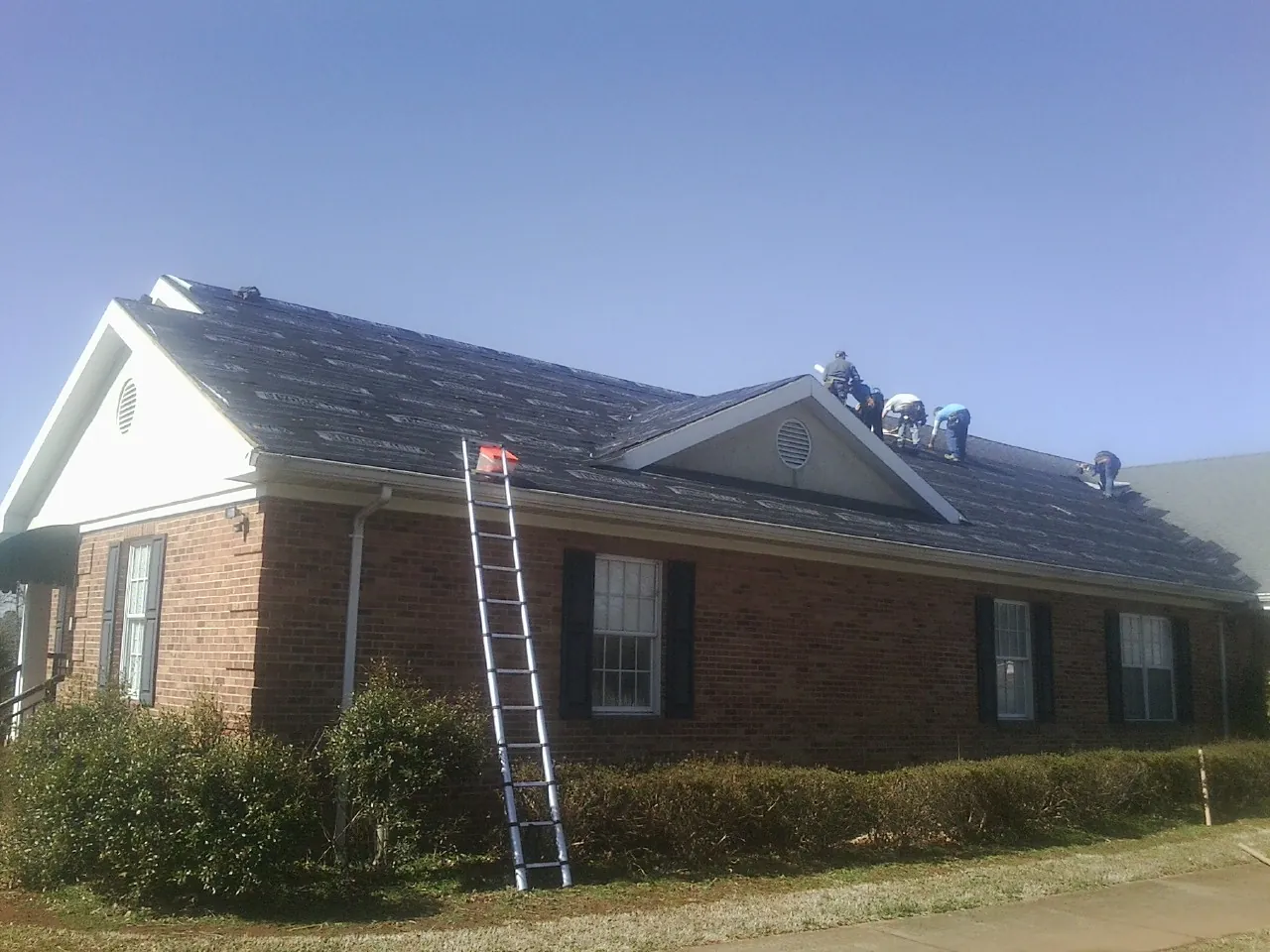 Crew tearing shingles off a brick ranch roof with a ladder set — Metal Roof Repair work by our crew in Woodlyn
