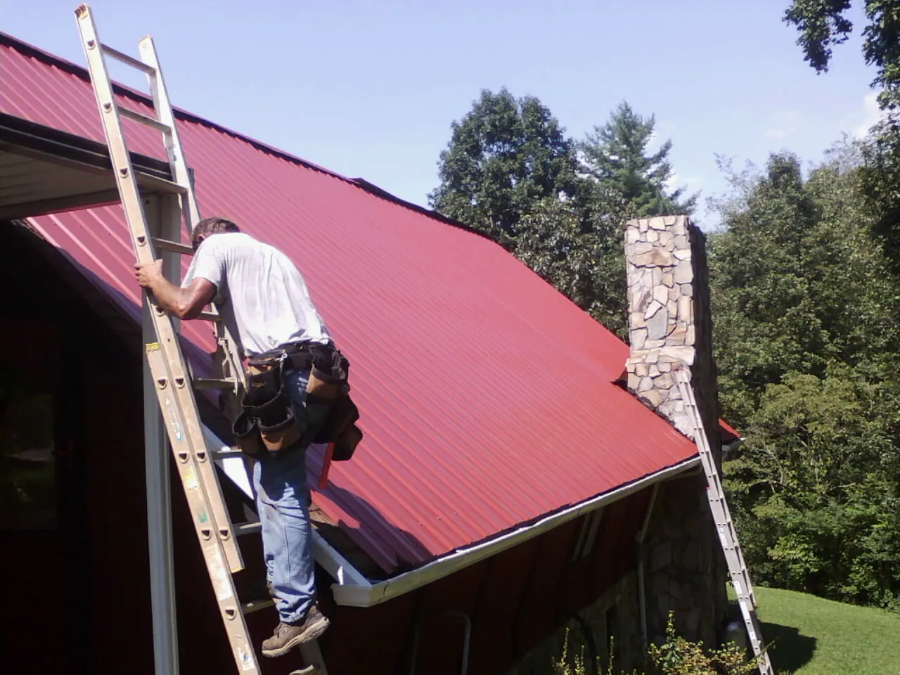 Roofer on a ladder beside a red metal roof and stone chimney — Soffit & Fascia Repair by Roofing Brothers in Woodlyn